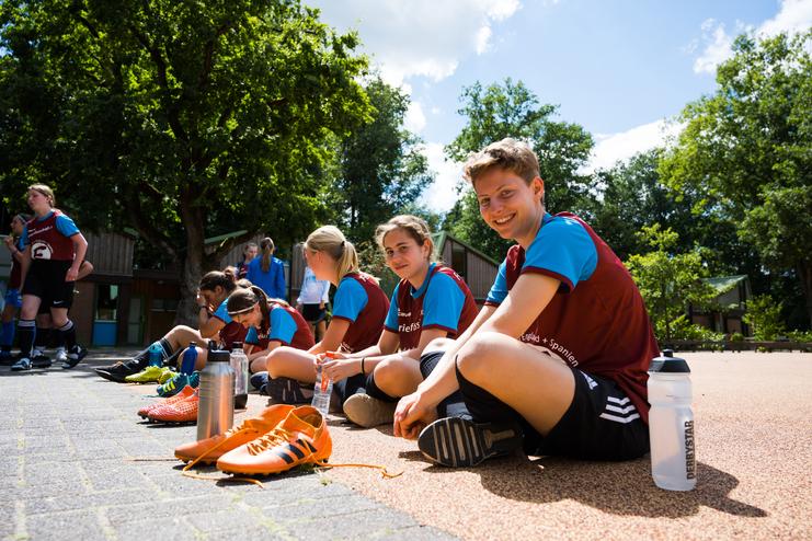 Eine Gruppe junger Fußballspielerinnen sitzt entspannt auf dem Boden, während sie ihre Schuhe ausziehen und sich unterhalten.